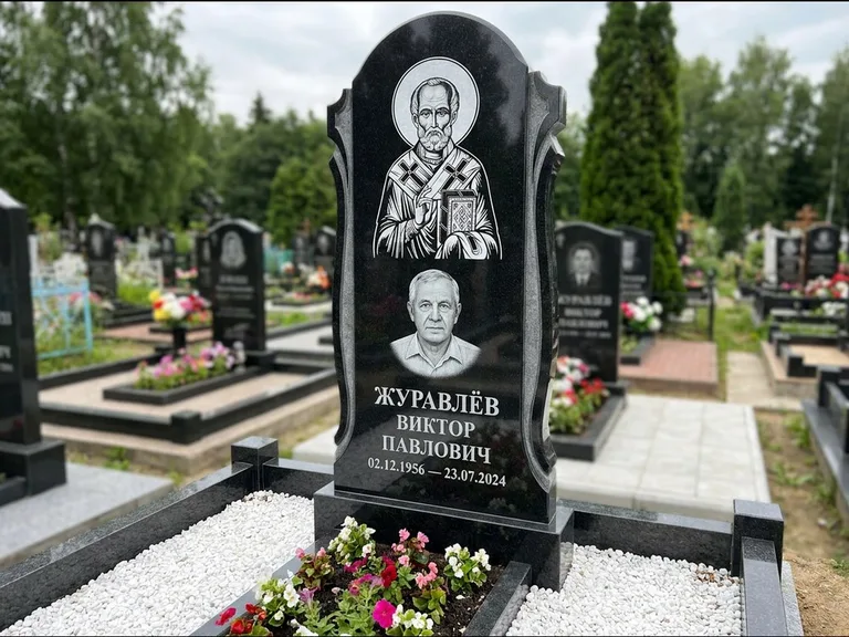 Памятник с гравировкой иконы: стела с образом в полный рост на обороте — гравировка образа Матроны Московской, низкие кусты спиреи в цвету, работа «Лебедева» в Челябинске